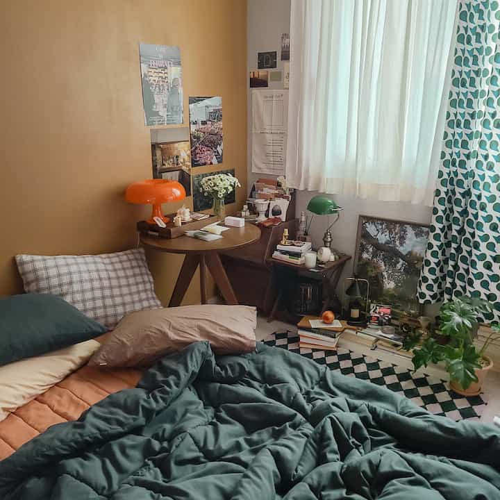 Cozy studio bedroom with warm orange wall and green bedding, featuring a round wooden table and vintage decor items
