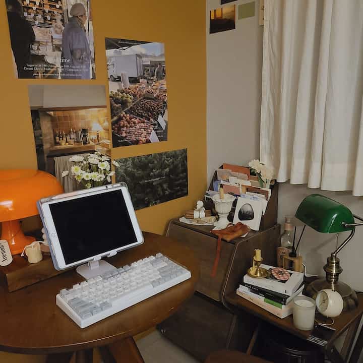 黄色の壁と木製の円形テーブルが配置されたワンルームのホームオフィス、ヴィンテージランプとポスターが印象的な空間