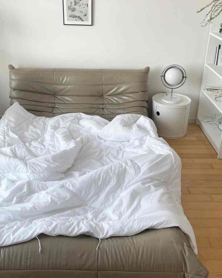 A clean studio bedroom in white and beige tones featuring a leather bed, white nightstand, and round table lamp