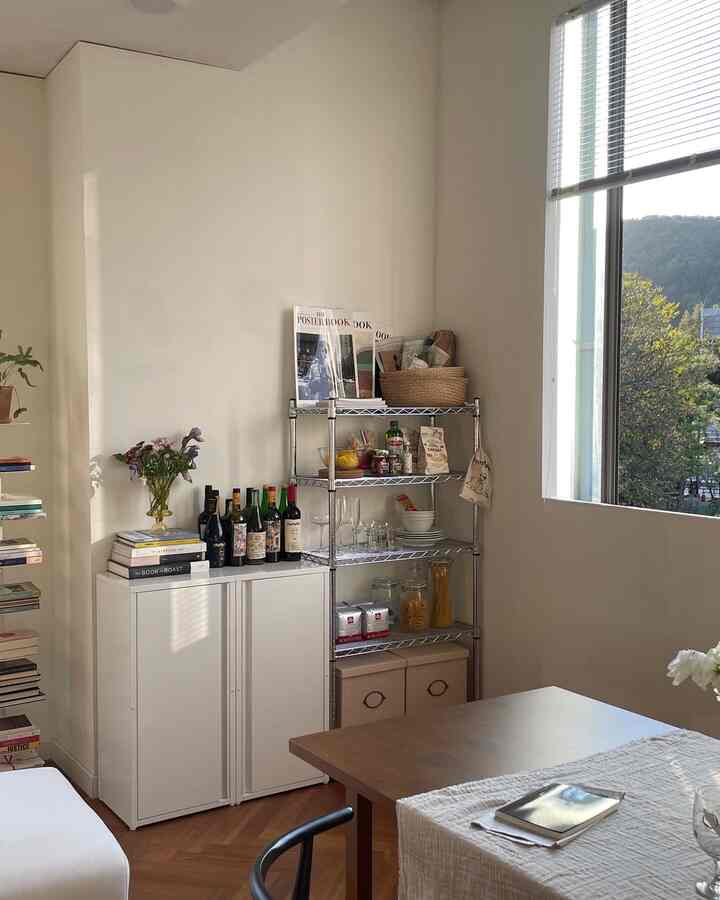 Natural-toned study corner with white walls, wood dining table, metal rack, and cabinets neatly arranged in warm ambiance