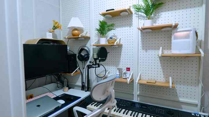 White-walled home office with wood-tone shelves, featuring a computer desk, microphone, and digital keyboard in a music workstation setup