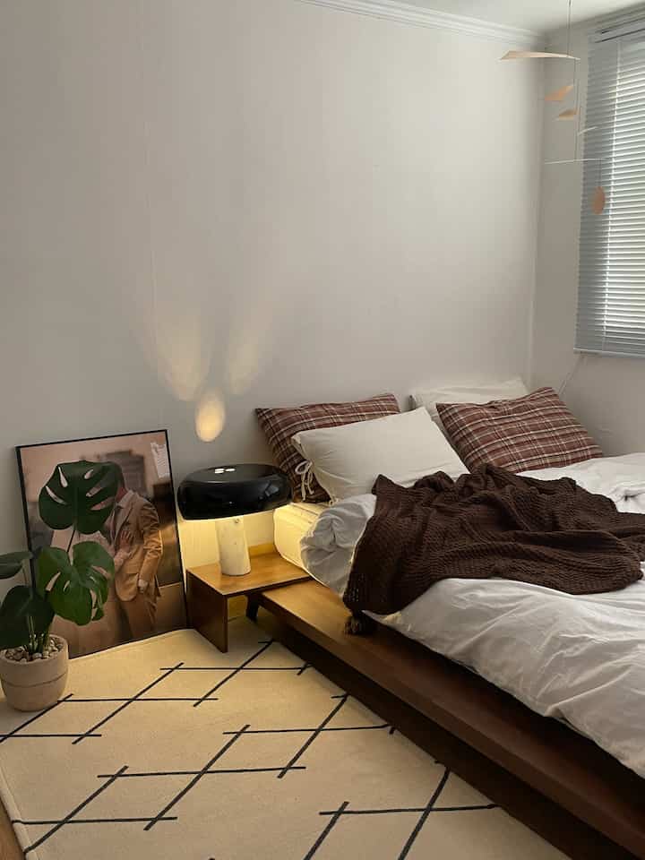 ナチュラルモダンなブラウンとホワイトの寝室、低めの木製ベッドとチェック柄クッション、テーブルランプが温かみのある雰囲気のあるインテリア