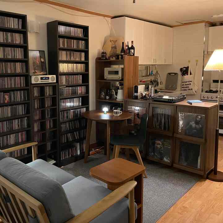 ナチュラルな木目調とグレーのソファがあるコンパクトなリビング、丸テーブルとCD棚が配置された雰囲気のあるホームカフェ空間