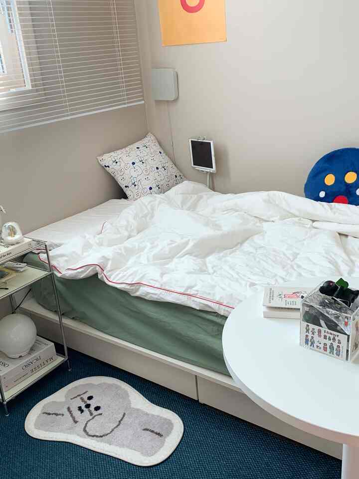 ホワイトとグリーンのトーンの小さな寝室、中央にマットレスカバーがかかったベッドと小さな円形テーブルが配置されたミニマルなインテリア