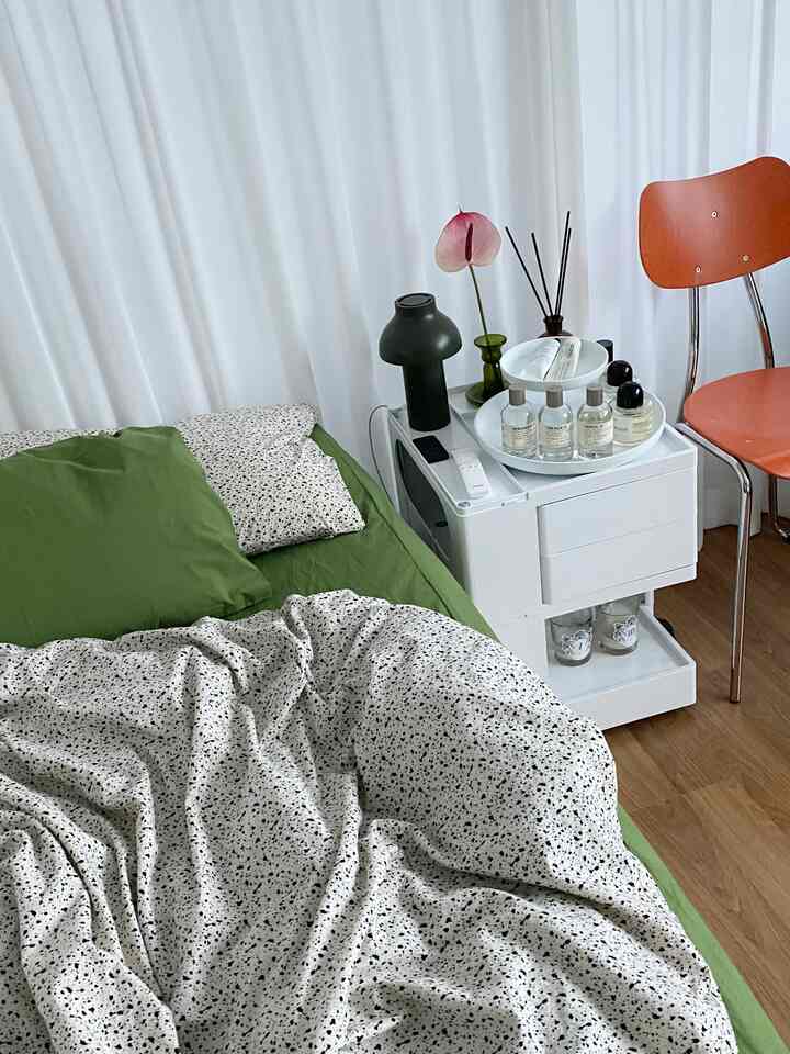Studio bedroom with white curtains and green bedding, featuring a modern design chair and white dresser in a cozy setting