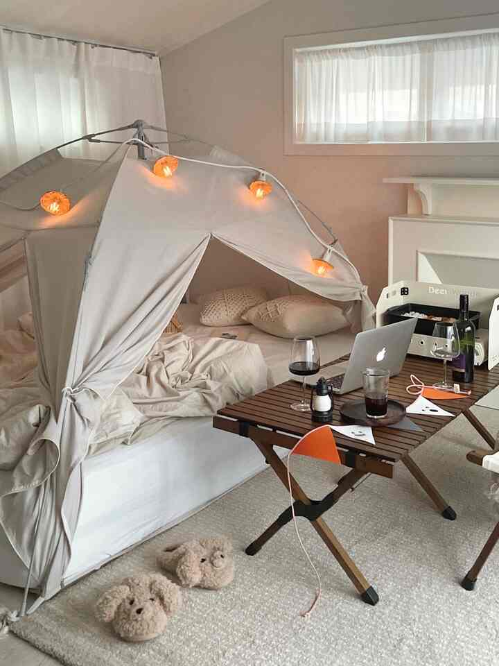 White and beige toned bedroom featuring a heating tent, camping table, and garland lights creating a cozy home camping atmosphere