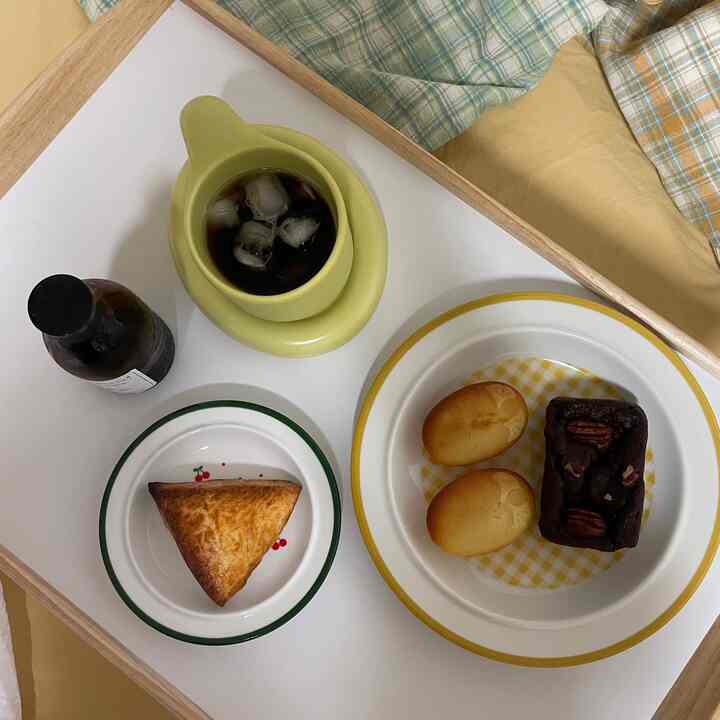 Soft yellow and natural wood-toned bed tray featuring iced coffee mug and plates with desserts in a cozy home cafe setting