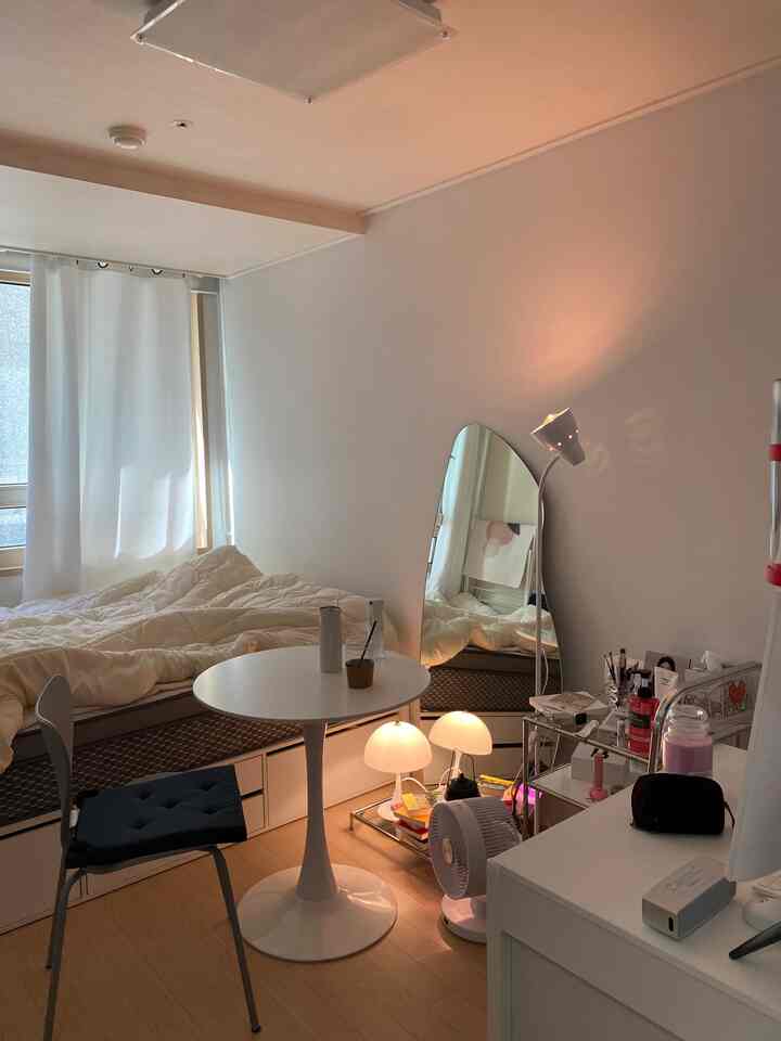 White and brown toned studio apartment featuring a bed, round table, full-length mirror, and floor lamp in a cozy modern interior