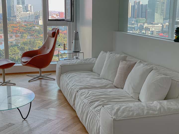Bright living room with white and brown tones featuring large windows, comfortable sofa, and armchair in a natural setting