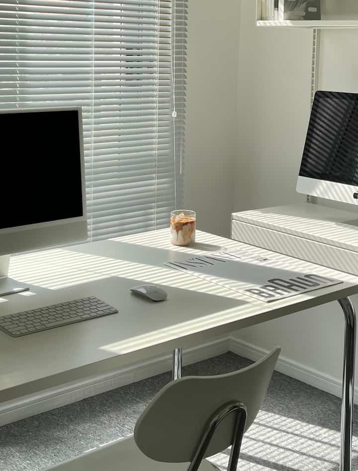 White toned study space featuring dual iMacs, aluminum blinds, and a coffee cup on a clean modern home office desk