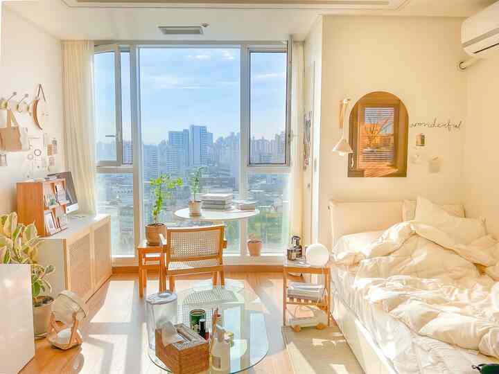 明るいベージュと木目調のワンルーム寝室、大きな窓とベッドリネン、木製のナイトテーブルと椅子が配置された自然な雰囲気のインテリア