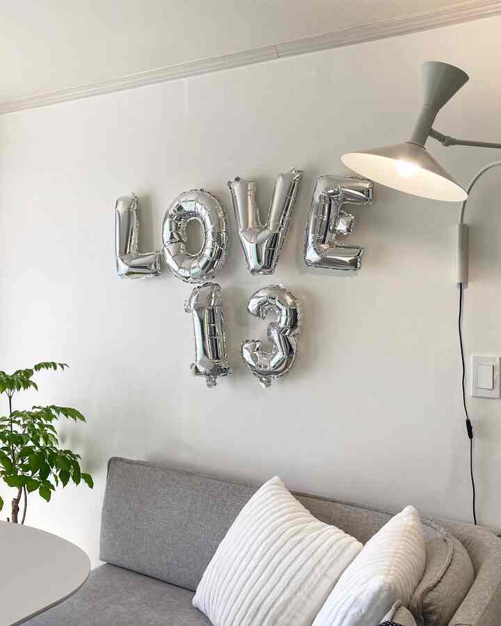 Bright white-walled living room with gray sofa, silver 'LOVE 13' balloons, and wall lamp creating a cozy atmosphere