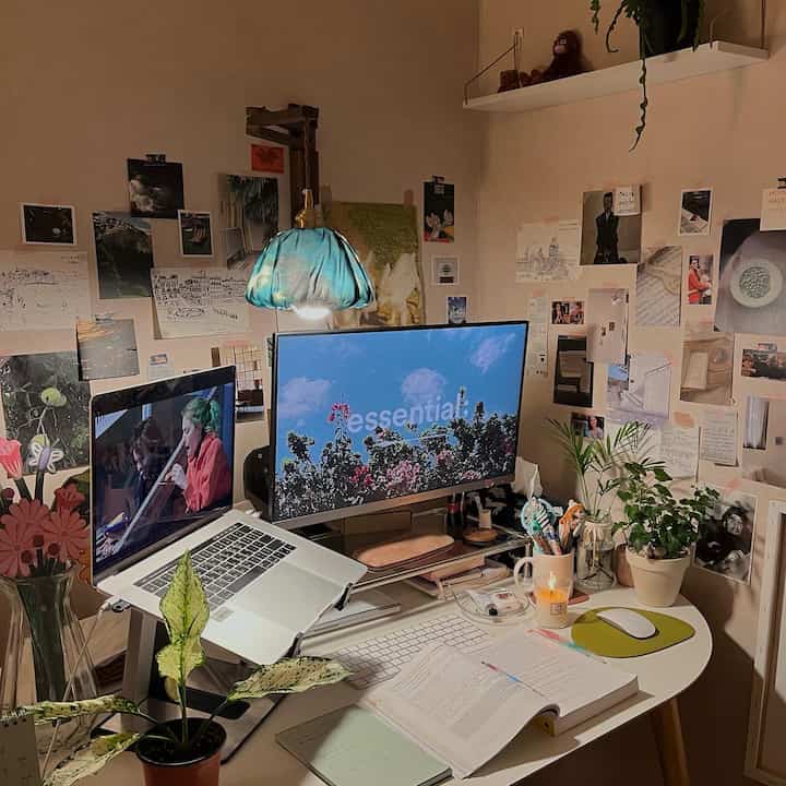 A white and wood tone home office featuring a desk with dual monitors, vintage posters on walls, and potted plants creating a cozy workspace