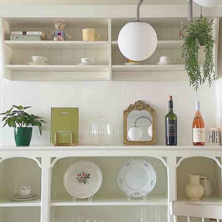 白を基調としたフレンチカントリー風キッチン、ヴィンテージの皿とアンティークな鏡が配置された整理された収納スペース