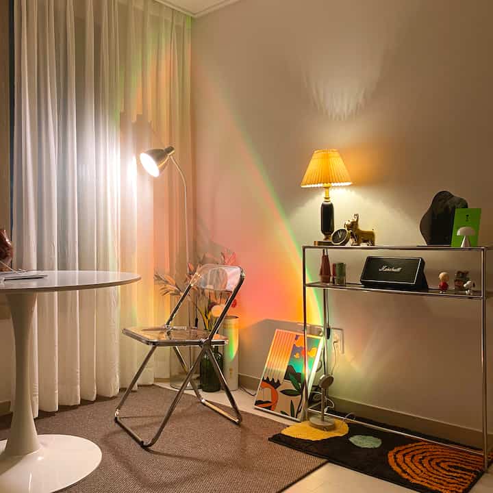 Warm-toned living room featuring white sheer curtains, transparent folding chair, round table, metal shelf, and various ambient lighting