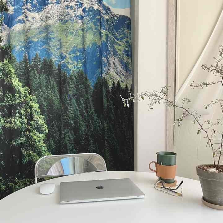 White-toned walls with a round dining table, transparent folding chair, and forest landscape curtain creating a Mid-Century Modern home office space