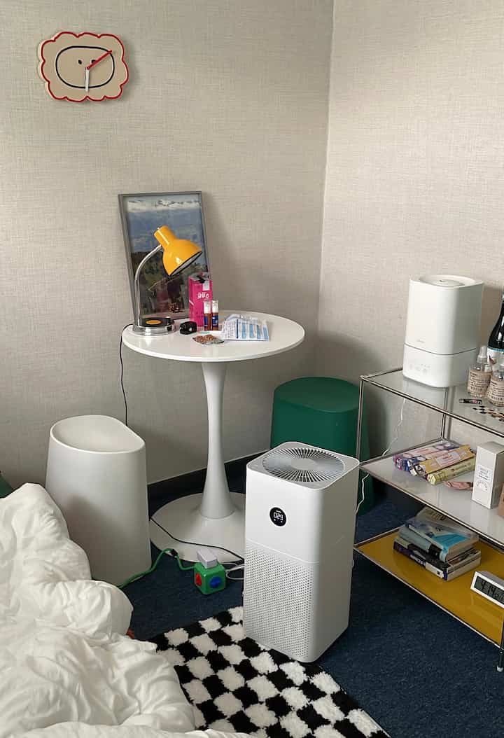 A small bedroom in white and blue tones featuring a mid-century style yellow table lamp, round dining table, and air purifier creating a cozy space