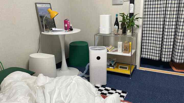 Compact bedroom in white and green tones featuring mid-century modern furniture, air purifiers, and a metal shelf with decor