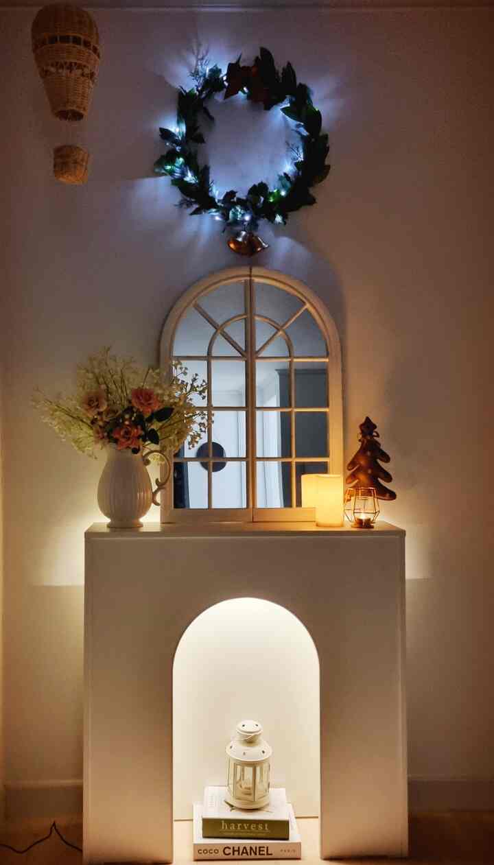 White and brown toned living room fireplace console featuring a Christmas wreath, arched mirror, vase, and mood lighting for cozy atmosphere