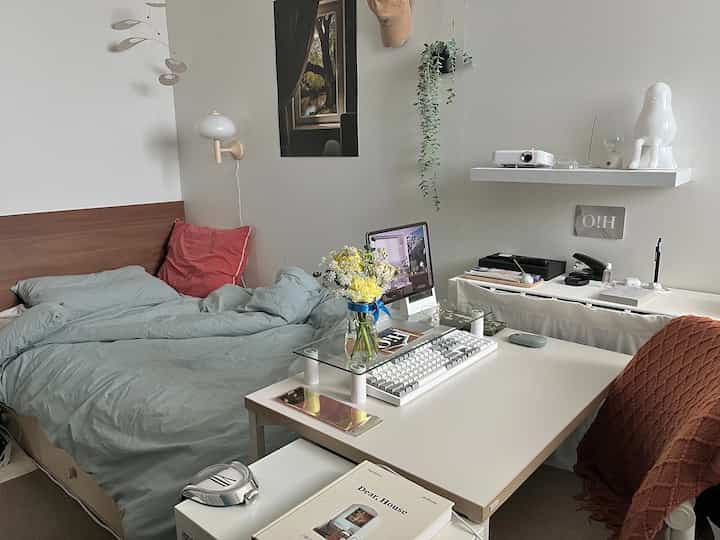 A cozy bedroom and home office space in white and gray tones, featuring a desk with a vase of flowers and a computer monitor