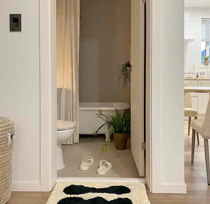 白とベージュのトーンのバスルームと隣接するキッチン、バスタブやバスマット、植物が配置された自然で落ち着いた雰囲気のインテリア