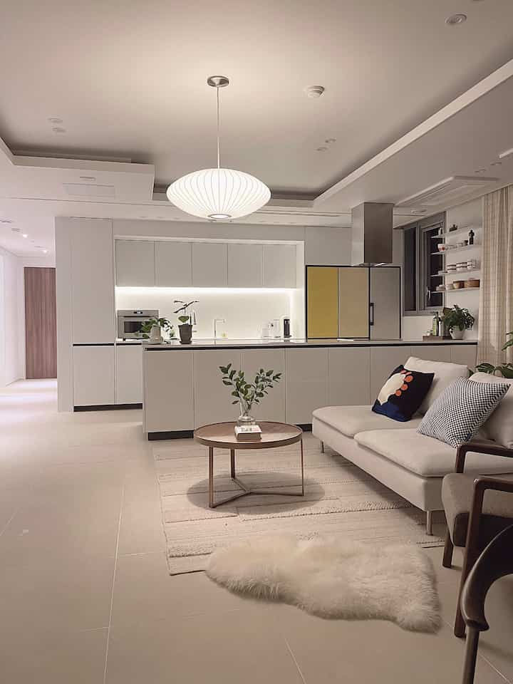 A clean living room and open kitchen in white and wood tones, featuring mid-century modern sofa and pendant lighting