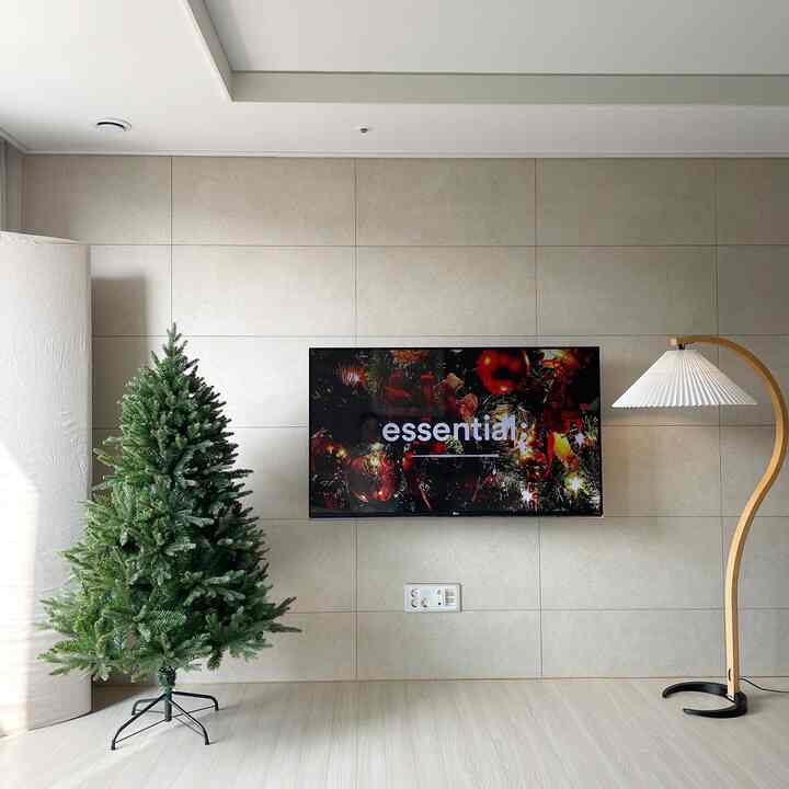 Beige tiled wall living room with wood-tone floor lamp, featuring a green Christmas tree on left and wall-mounted TV in center with a clean simple atmosphere