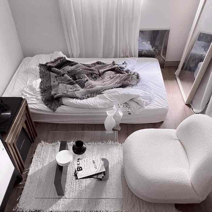 White and natural toned studio bedroom featuring a simple bed, rug, curtains, and a cozy armchair in a minimalist space