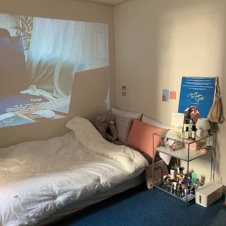 白い寝具と青いラグが目立つワンルームの寝室、壁には映画のポスターとプロジェクターの映像が映る雰囲気のあるインテリア