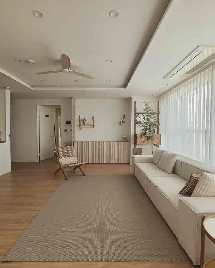 Beige and wood tone living room featuring a beige sofa, wooden chair, large window with sheer curtains creating a warm and simple atmosphere.