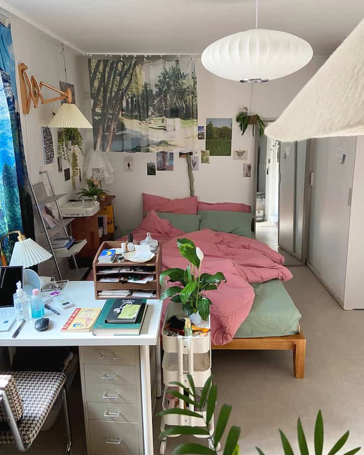 White-toned studio bedroom featuring pink and green bedding, a desk, and various plants creating a natural atmosphere