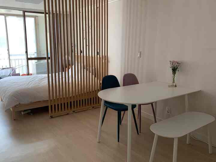 A modern natural studio apartment bedroom with white and wood tones featuring a dining table, chairs, and a wooden room divider