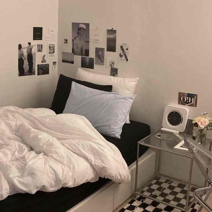 Modern bedroom in white and black tones featuring a bed with layered pillows, posters on the wall, and a transparent nightstand with decor items