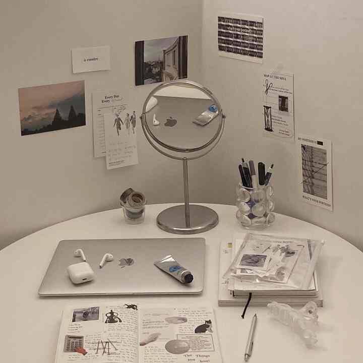Monotone white-toned home office space featuring a round table with laptop, reading materials, a mirror, and posters arranged neatly for a clean workspace