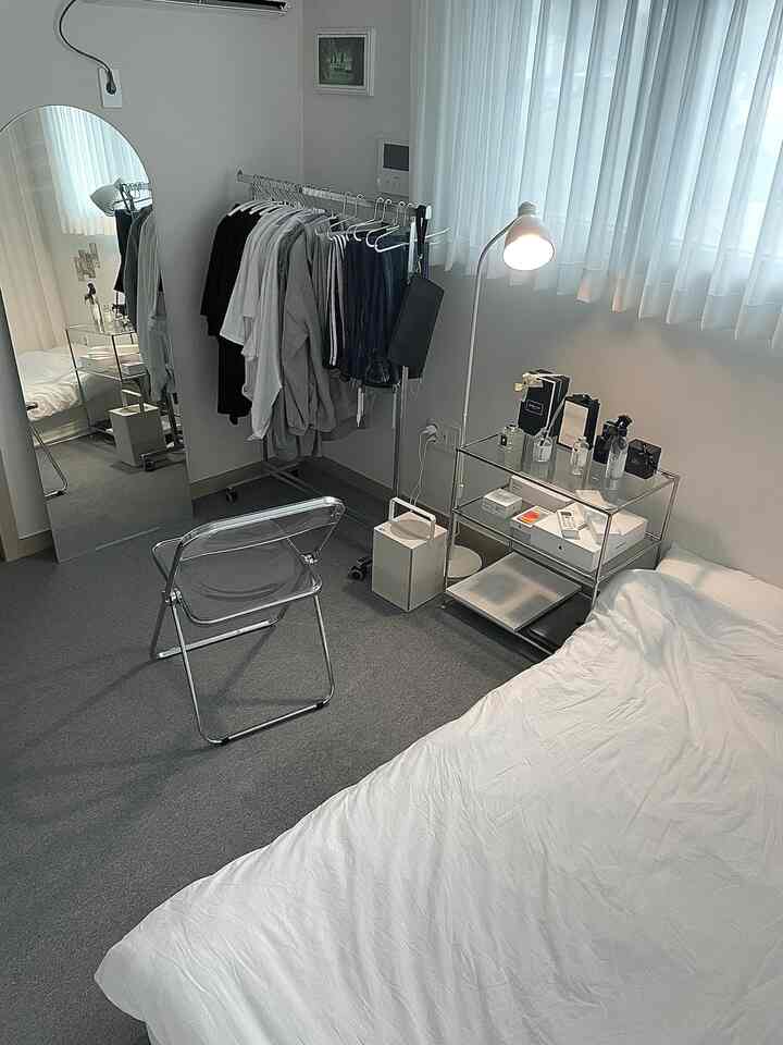 白とグレーのトーンの寝室、モダンなミニマリストスタイルで椅子、全身鏡、カーテンが配置された居心地の良い一人暮らしの部屋