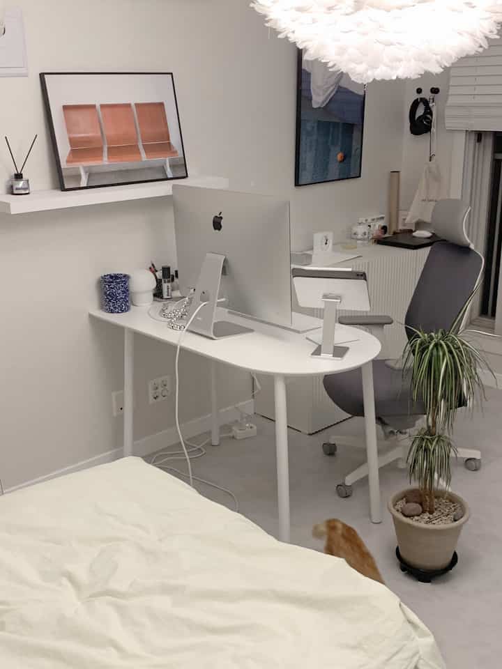 A modern, compact home office in white tones featuring a desk, ergonomic chair, and a potted plant with an orange cat on the floor.