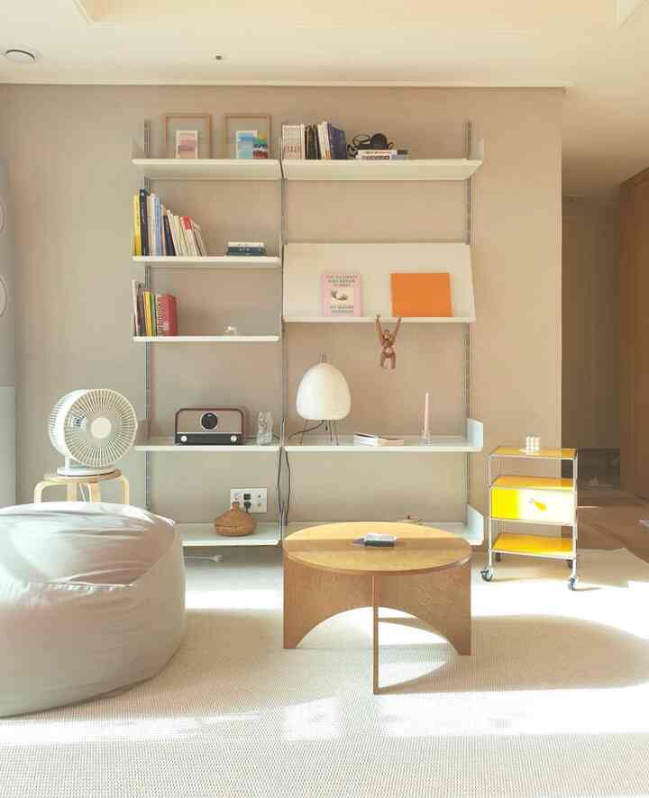 明るいベージュと白の色調のリビングルーム、木製のコーヒーテーブルと白いビーンバッグ、壁に取り付けられた棚と小さなインテリア小物が配置された落ち着いた清潔感のある空間