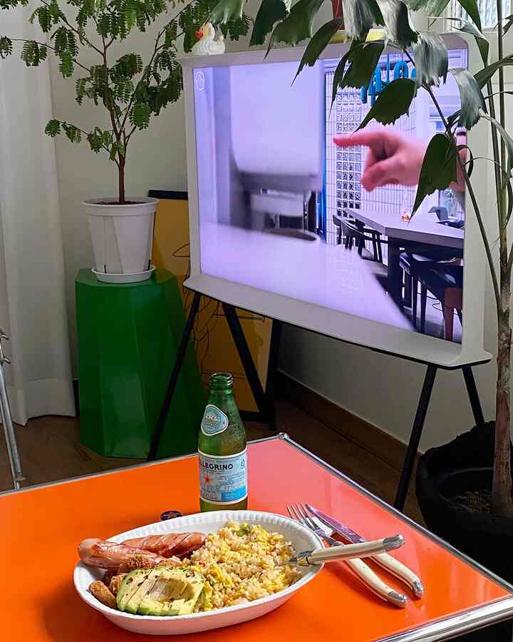 White and green toned studio living room featuring an orange dining table with plate and cutlery, and a Samsung Serif TV in a modern setting