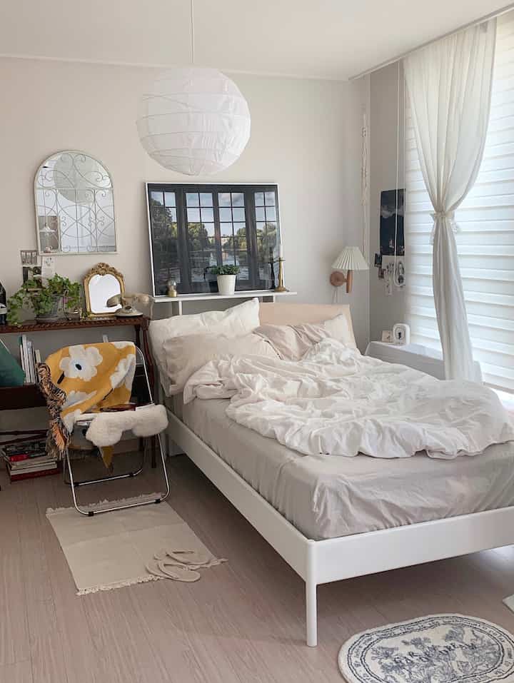 Soft white and natural toned bedroom featuring a neat bed frame, curtains, vintage decor, and plants creating a cozy atmosphere