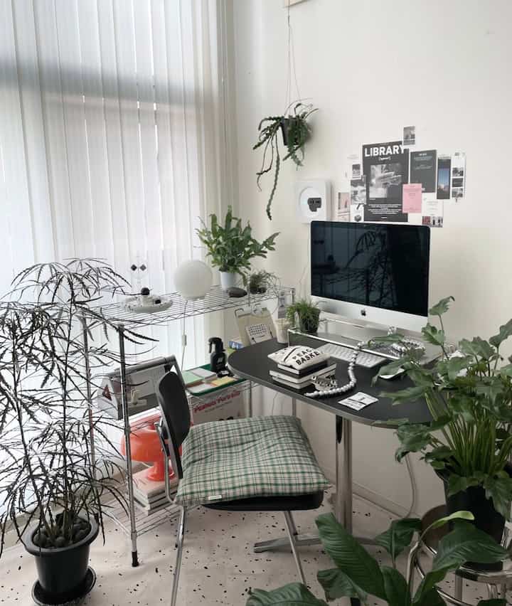 Bright white-walled studio with black metal shelf, desk, and multiple green plants creating a cozy home office