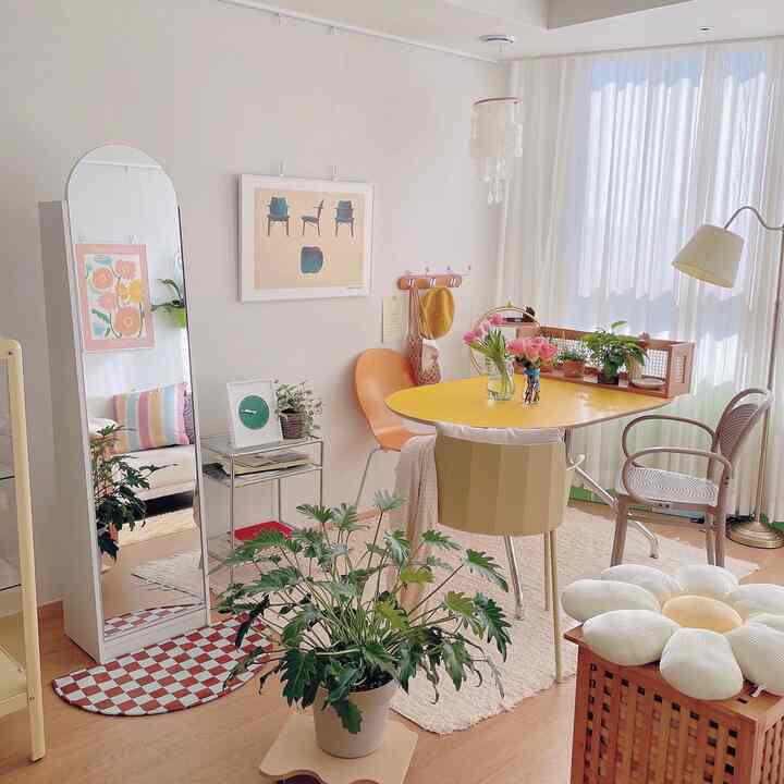 White and yellow toned studio apartment living room featuring a round full-length mirror, dining table, and colorful chairs in a cozy natural setting