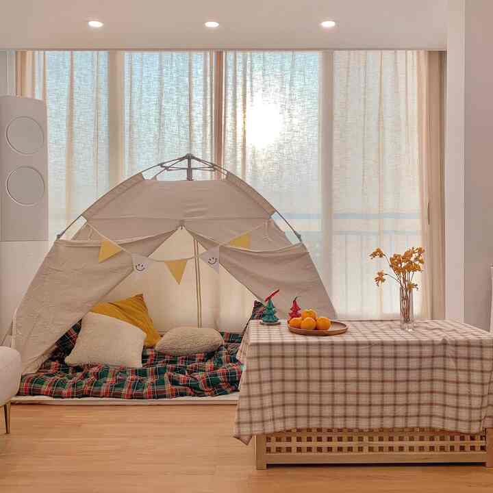 A cozy living room with beige linen curtains and warm wood tones, featuring a heating tent with cushions at center for home camping