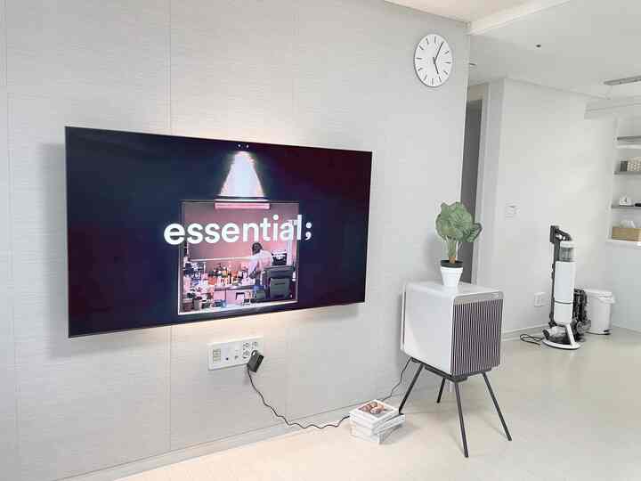 White and natural tone living room featuring a wall clock, wall-mounted TV, air purifier on stand, and cordless vacuum for a clean, modern space