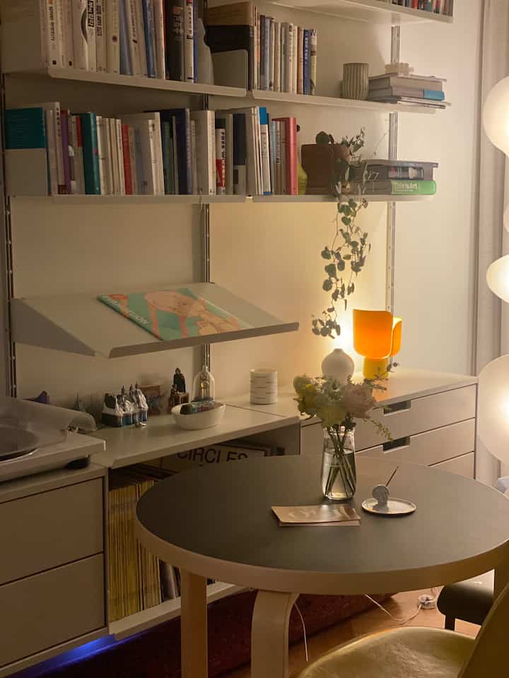 Beige and black toned living room featuring a round table, shelves with flowers, and warm lighting creating a cozy atmosphere