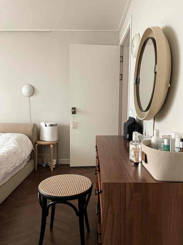 Natural wood tones and white bedroom featuring wooden vanity, round mirror, and rattan stool creating calm atmosphere