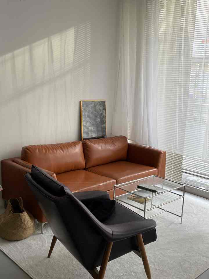 白い壁とカーテン、茶色のレザーソファ3人掛け、黒のアームチェア、ガラスのコーヒーテーブルが配置された自然な雰囲気のオフィス隣の休憩スペース