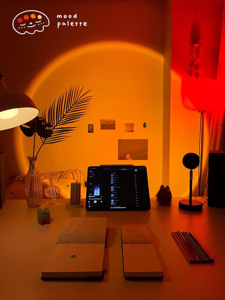 暖かいオレンジと赤のムード照明が照らすワンルームのデスク空間、ノートとタブレットが置かれた居心地の良い個人作業スペース