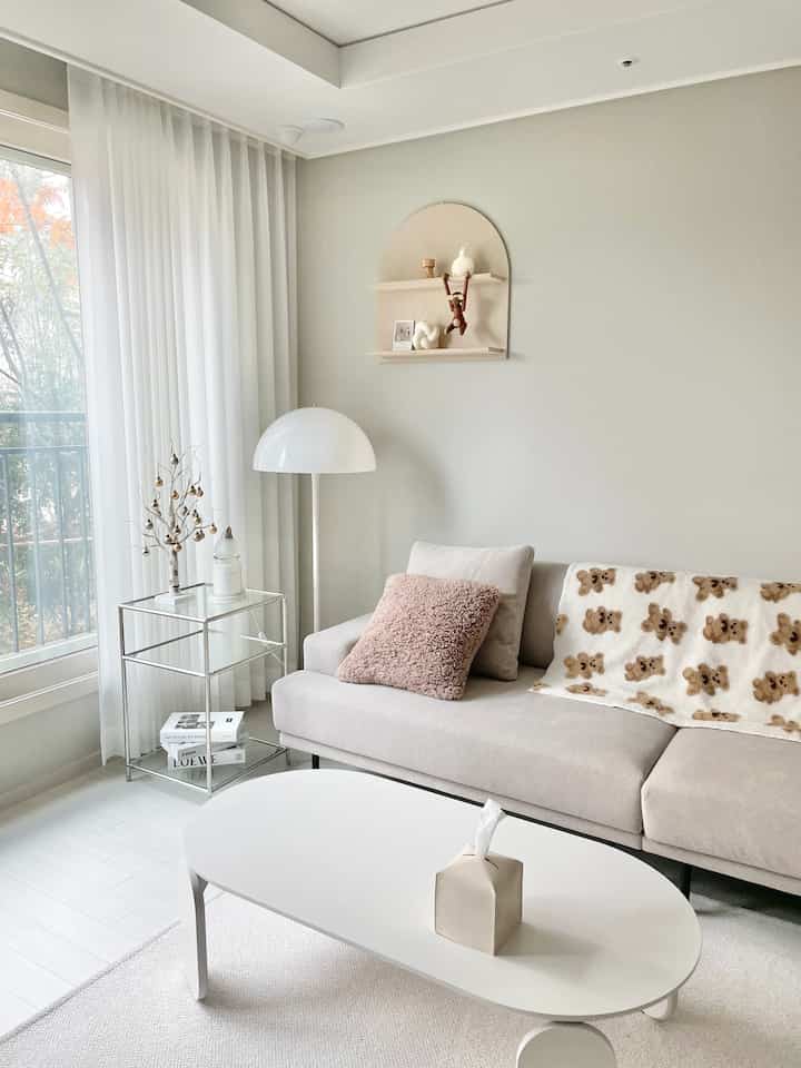 A white and beige toned living room featuring a beige sofa, white coffee table, floor lamp, and decorative objects on a wall shelf, creating a cozy atmosphere