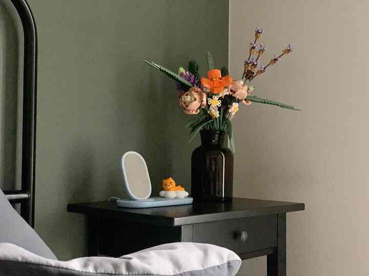 A natural bedroom scene featuring a dark brown and black nightstand with a brown glass vase and a LEGO flower bouquet on top