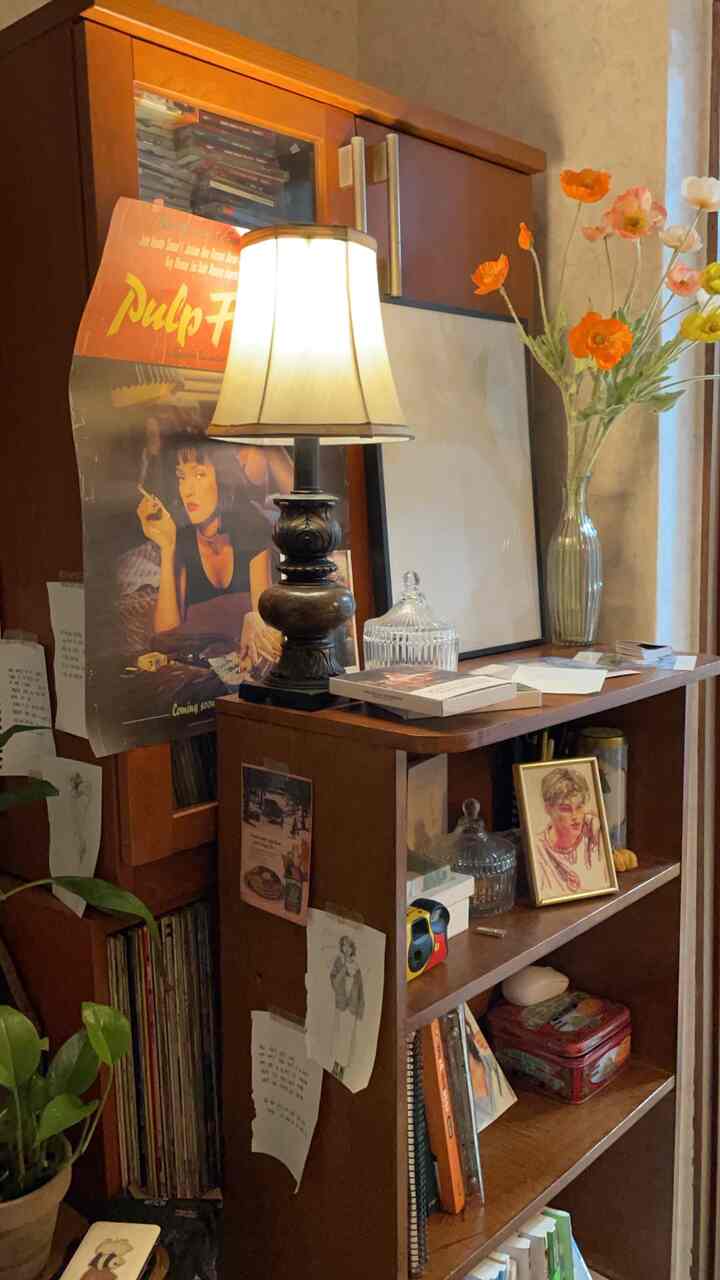 A warm natural-toned study space featuring a wooden bookshelf, antique table lamp, and a vase with poppy artificial flowers.